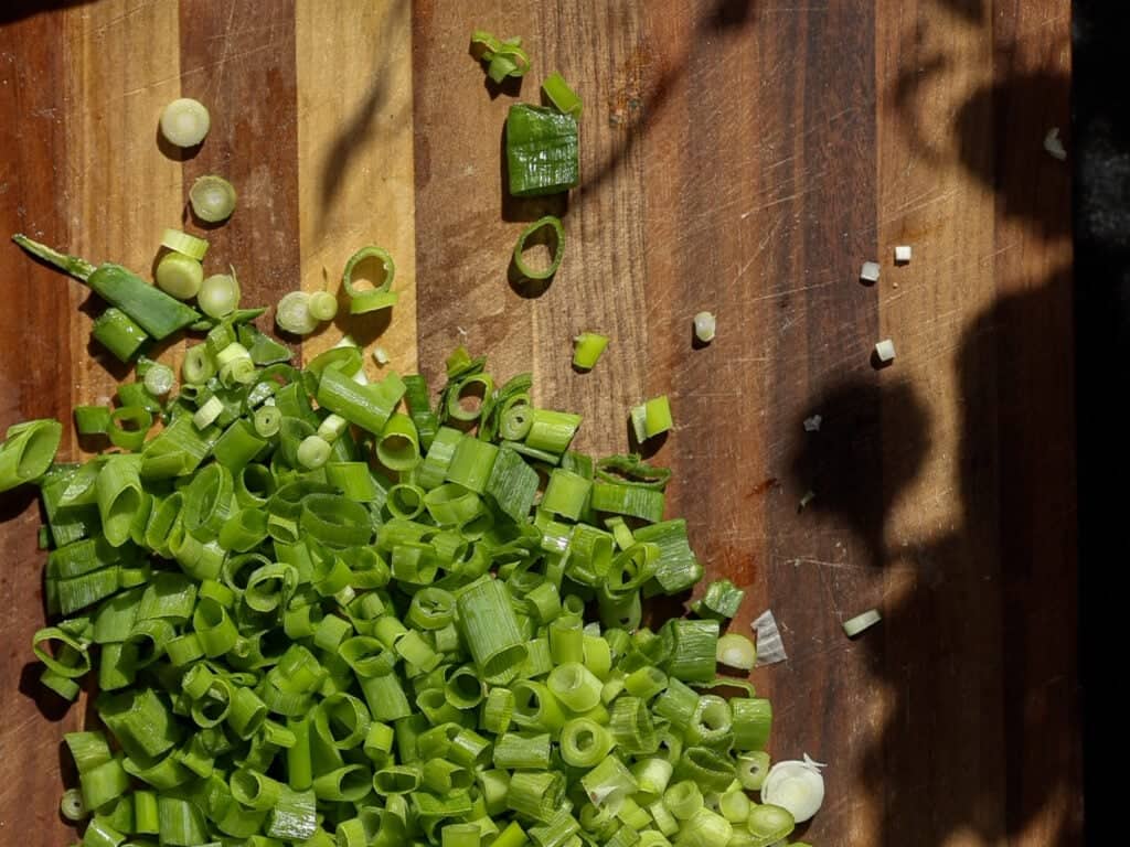 Chopped green onions scattered on a wooden cutting board, with some shadows cast across the surface.